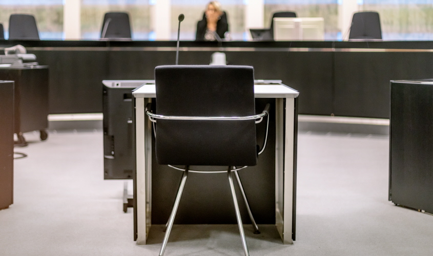 Bild på en förhandlingssal. En tom stol och i bakgrunden ser man en oskarp bild på en domare. Foto. 