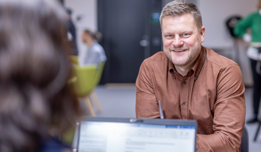 Man tittar in i kameran, sitter mittemot en person med laptop.