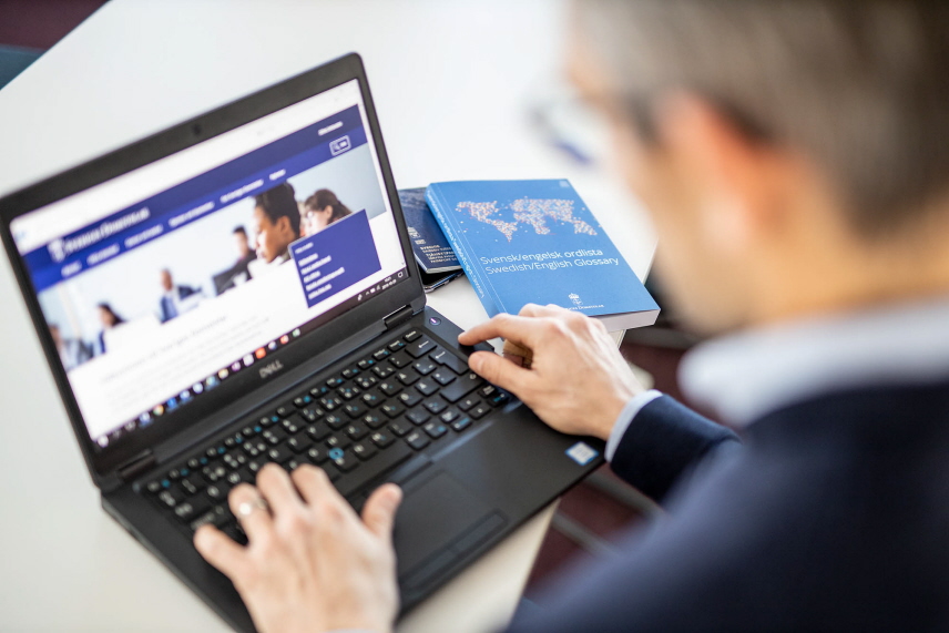 En person sitter och arbetar vid sin bärbara dator. Bredvid sig har han en engelsk-svensk ordbok, pass och telefon.