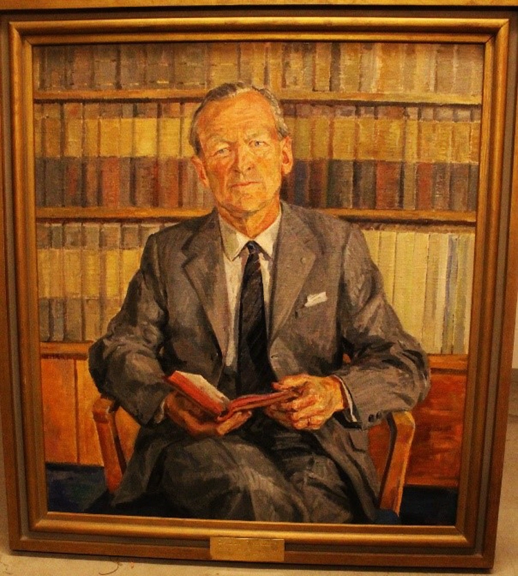 Inramat porträtt av medelålders lätt gråhårig man i grå kostym med uppslagen bok i handen, sittandes framför välfyllda bokhyllor