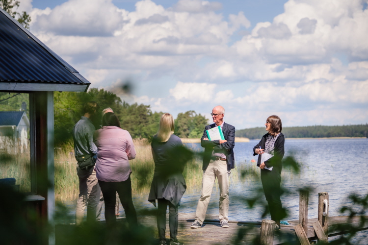 Fem personer som står vid en sjö och diskuterar. Foto.