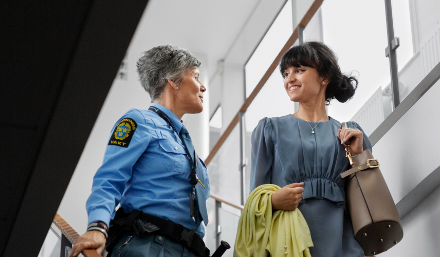 Två kvinnor går nedför en trappa och pratar med varandra. Den ena arbetar som ordningsvakt och har en blå uniform på sig.