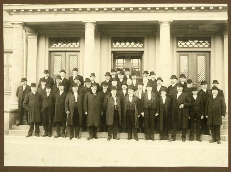 En grupp män klädda i tidsenliga rockar och hattar ståendes på tingshusets trappa, svartvitt