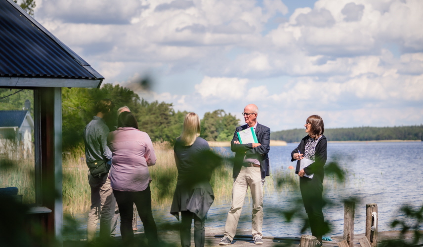 Fem personer som står vid en sjö och diskuterar. Foto.